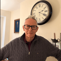 Chris Swithinbank stands indoors in front of a large wall clock, wearing a grey cable‑knit jumper